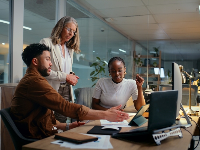 Three colleagues reviewing financial reports and charts around a desk in a modern office at night