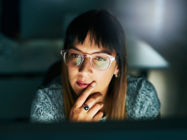 Woman in glasses studying a screen in a dark office, focused on analyzing customer feedback