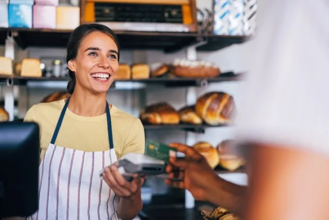 A smiling baker in a striped apron holds a payment terminal as a customer pays with a card, representing a successful transaction and customer satisfaction.