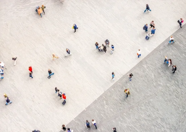 Aerial view of people walking on a large, light-colored paved plaza divided by a diagonal line into two slightly different shades of gray paving stones.