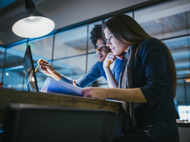 Two colleagues reviewing work on a laptop under a lamp in a dark office at night