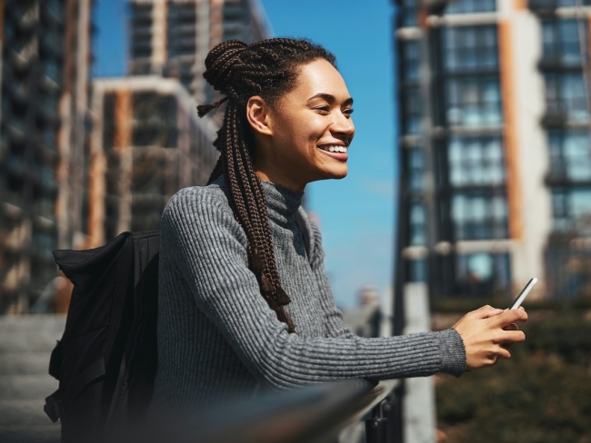 Young woman smiling while using a smartphone outdoors near urban buildings, representing citizens accessing government digital services.