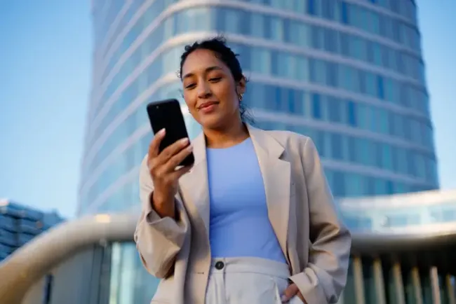 Smiling businesswoman using a smartphone in front of a sleek, modern office building.