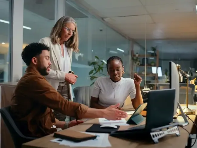Three colleagues reviewing financial reports and charts around a desk in a modern office at night.
