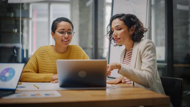 Two women collaborating over documents and a laptop at a wooden table in an office setting.