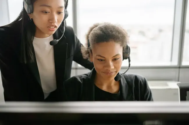 A supervisor standing over a customer service representative, pointing to something on a computer screen. They are in a call center setting, appearing to be in a customer service skills training or coaching session.