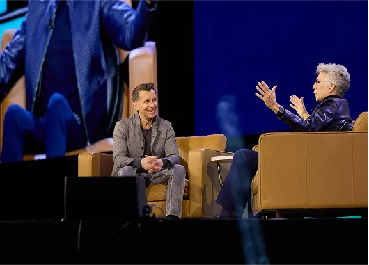 Two business professionals in conversation on stage with tan leather chairs against blue backdrop