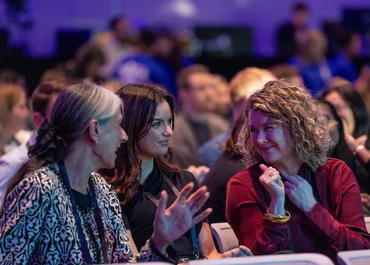 Three women engaged in conversation at conference table with purple lighting and blurred audience in background